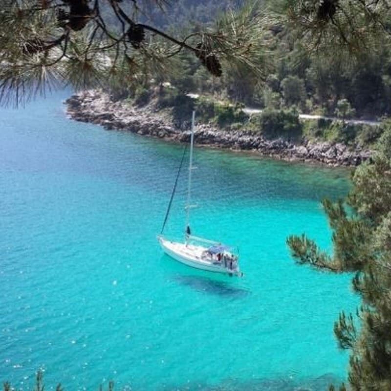 A sailing yacht at anchor in a turquoise cove under pine trees