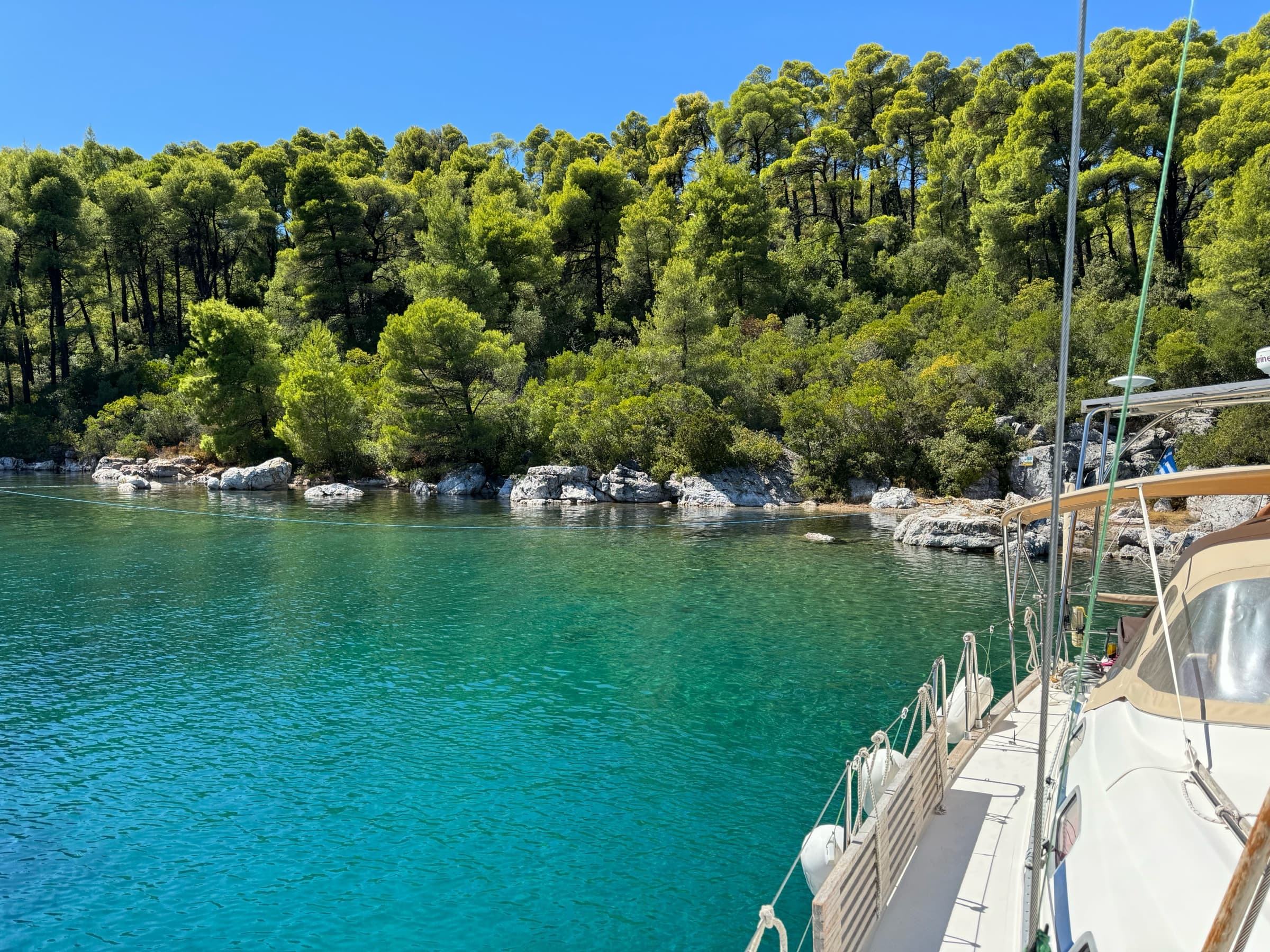 The yacht at anchor in a turquoise cove lined with pine trees